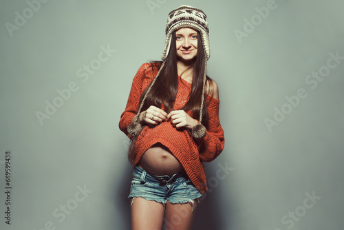 Stylish pregnancy concept. Portrait of happy hipster mommy with hands on stomach posing in trendy knitted clothing over light gray background. Urban street style. Copy-space. Studio shot