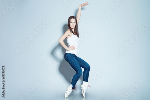 Dancing Queen concept. Full length portrait of young fitness model in white blank sleeveless shirt and blue jeans dancing with hand up over light blue background. Copy-space. Studio shot photo