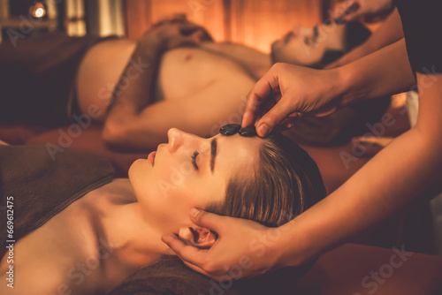 Caucasian couple enjoying relaxing anti-stress head massage with hot stone and pampering facial beauty skin recreation leisure in warm candle light ambient salon spa in resort or hotel. Quiescent