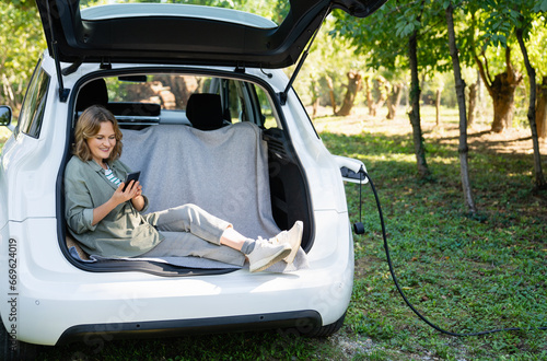 Woman with smartphone sits in an electric car's trunk © scharfsinn86