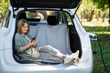 Woman with smartphone sits in an electric car's trunk