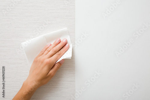 Young adult woman hand holding white dry paper napkin and wiping surface from glue after light gray wallpaper gluing. Closeup. Front view. Repair work of home.