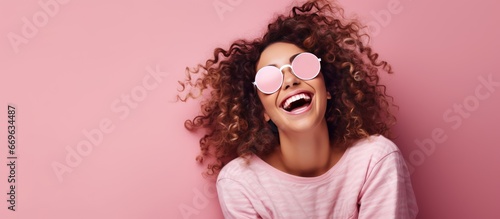 Joyful young woman with tattoos curly hair and sleep mask happily hopping while holding a pillow over a pink background photo