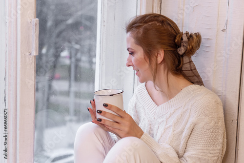 Close-up portraits of young stylish woman sitting on windowsill, drinking cocoa from cup, looking out window. Winter festive atmosphere. Girl in home clothes drinks tea, coffee. Holiday light bulbs.