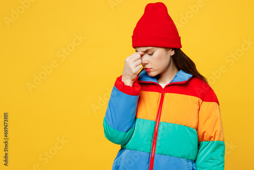 Young sad sick ill woman tired she wears padded windbreaker jacket red hat casual clothes keep eyes closed rub put hand on nose isolated on plain yellow background studio portrait. Lifestyle concept.