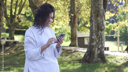 Young brunette girl in white sweatshirt typing on her mobile phone and taking a smiling selfie in a natural green setting