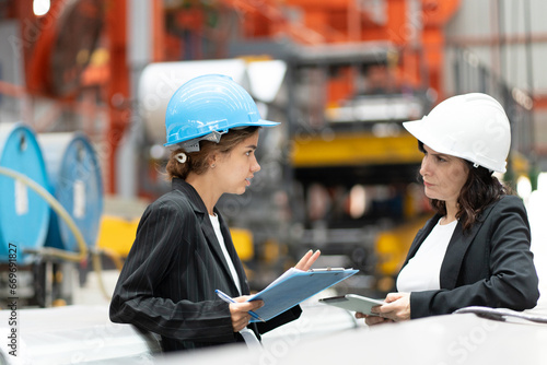 two professional woman engineer,worker,technician use clipboard discuss work, walk in steel metal manufacture factory plant industry. woman wear hard hat check quality machine