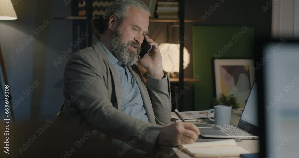 Businessman making mobile phone call and taking notes writing in notebook in office in evening. Business communication and night shift job concept. Businessman making mobile phone call and taking notes writing in notebook in office in evening. Business communication and night shift job concept.