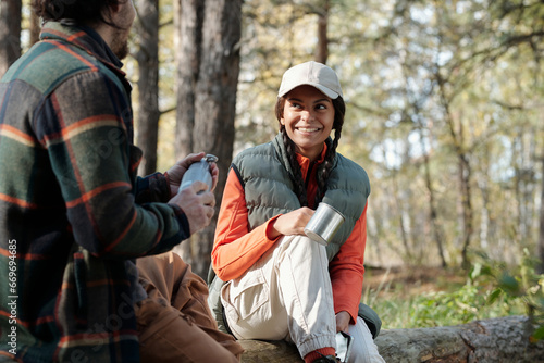 Young cheerful African American woman with metallic mug looking at her husband or boyfriend while sitting in front of him in the forest