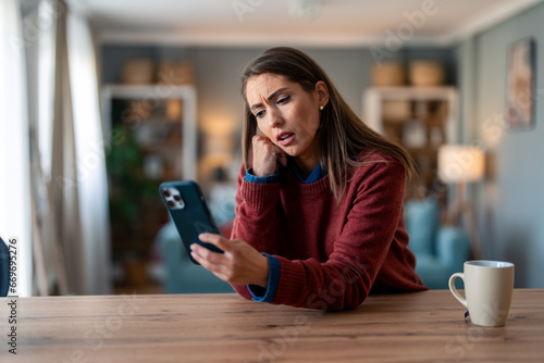 Stressed businesswoman with hand on cheek looking at smart phone while sitting at desk at home.