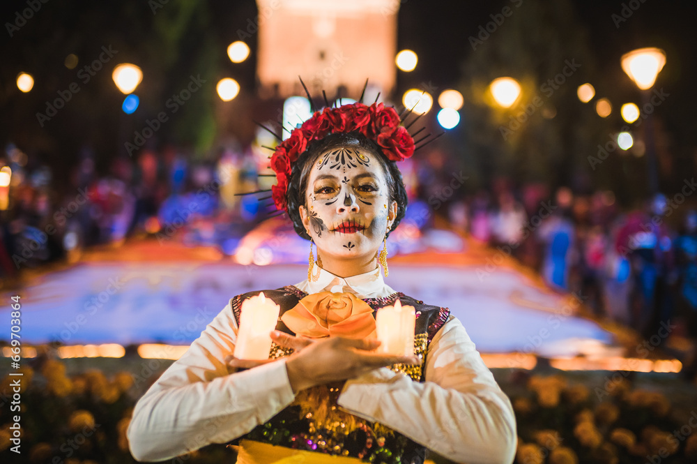 Mexican woman dressed and characterized as a catrina for the Day of the Dead festivity, with cempasuchil flowers and candles, behind her an offering of the dead.