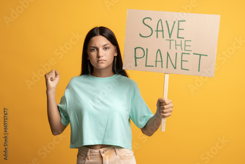 Serious confident brunette teen girl eco activist showing holding save the planet poster on yellow background looking at camera. Environmental protection, defend nature, picket demonstration concept. photo