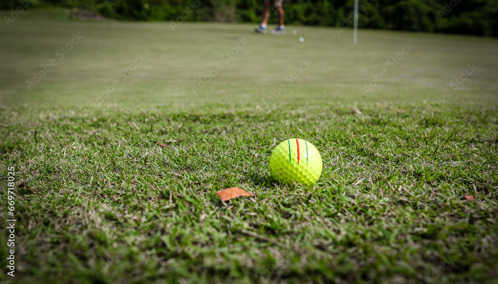 Yellow striped golf ball next to brown leaf on the fringe of a golf green with golfer and hole in background in Central Florida