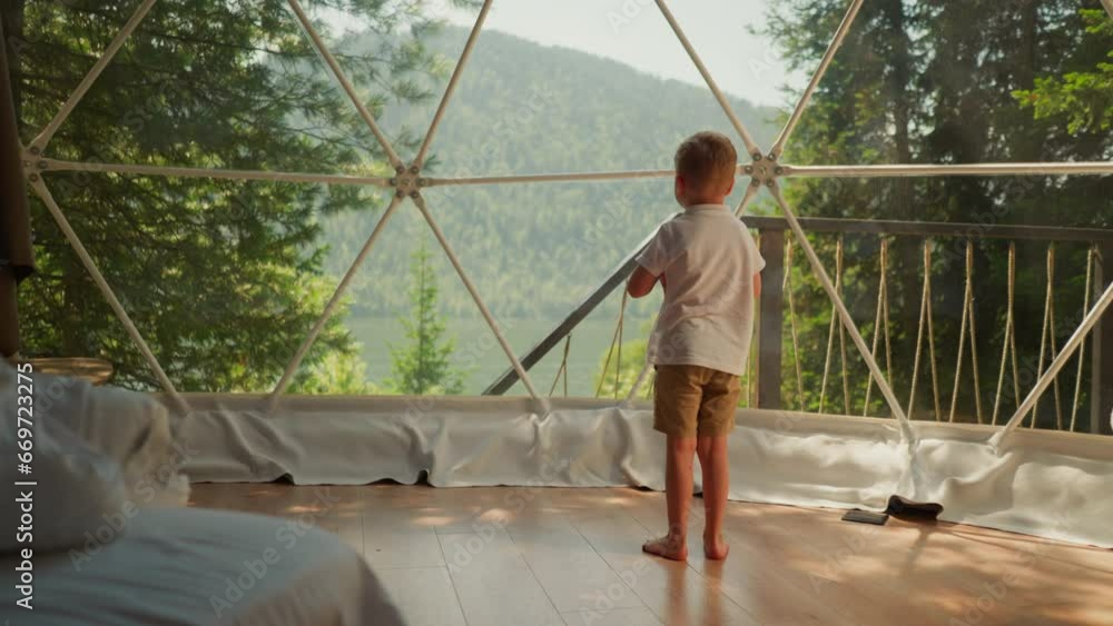 Child stands alone under glass dome at glamping. Thoughtful little boy looks out of panoramic window on terrace. Kid observes calm nature scene Child stands alone under glass dome at glamping. Thoughtful little boy looks out of panoramic window on terrace. Kid observes calm nature scene