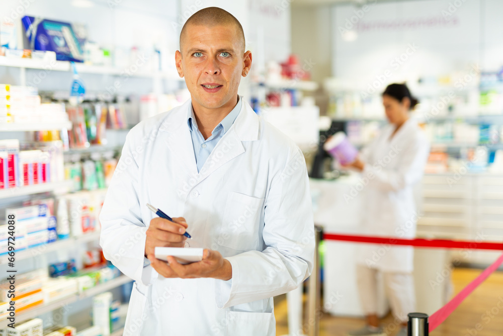 Male pharmacist with pan and paper in hands standing in drugstore and looking in camera. His female colleague working in background.