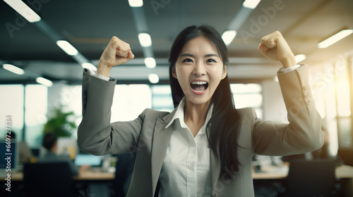 Cool looking young Asian businesswoman raising her fists in success and joy in the office. © Tepsarit