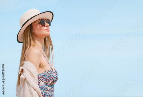 Young woman in a swimsuit, shirt, sunglasses and hat posing gracefully on a sandy beach against a blue sky