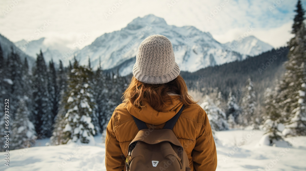 Fototapeta premium Rear view of a young traveler girl in a hat standing over snow-covered mountain peaks. Winter travel scene, wanderlust concept