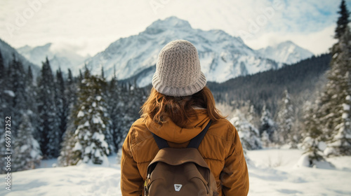 Rear view of a young traveler girl in a hat standing over snow-covered mountain peaks. Winter travel scene, wanderlust concept