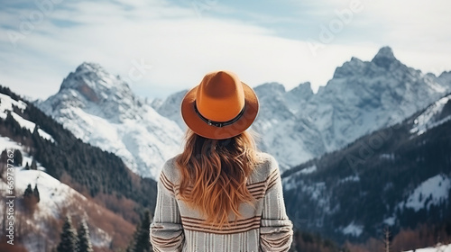 Rear view of a young traveler girl in a hat standing over snow-covered mountain peaks. Winter travel scene, wanderlust concept