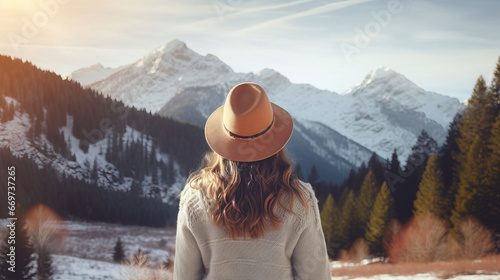 Rear view of a young traveler girl in a hat standing over snow-covered mountain peaks. Winter travel scene, wanderlust concept #669737265