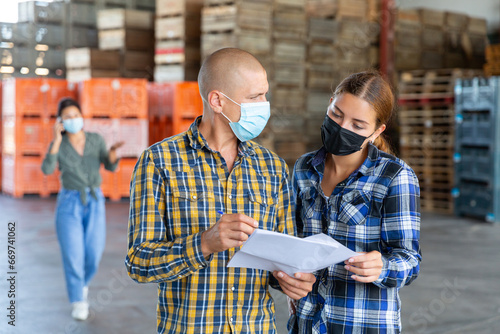 Man and young woman in face masks talking about documentation while standing in warehouse. Another woman talkin on phone in background.