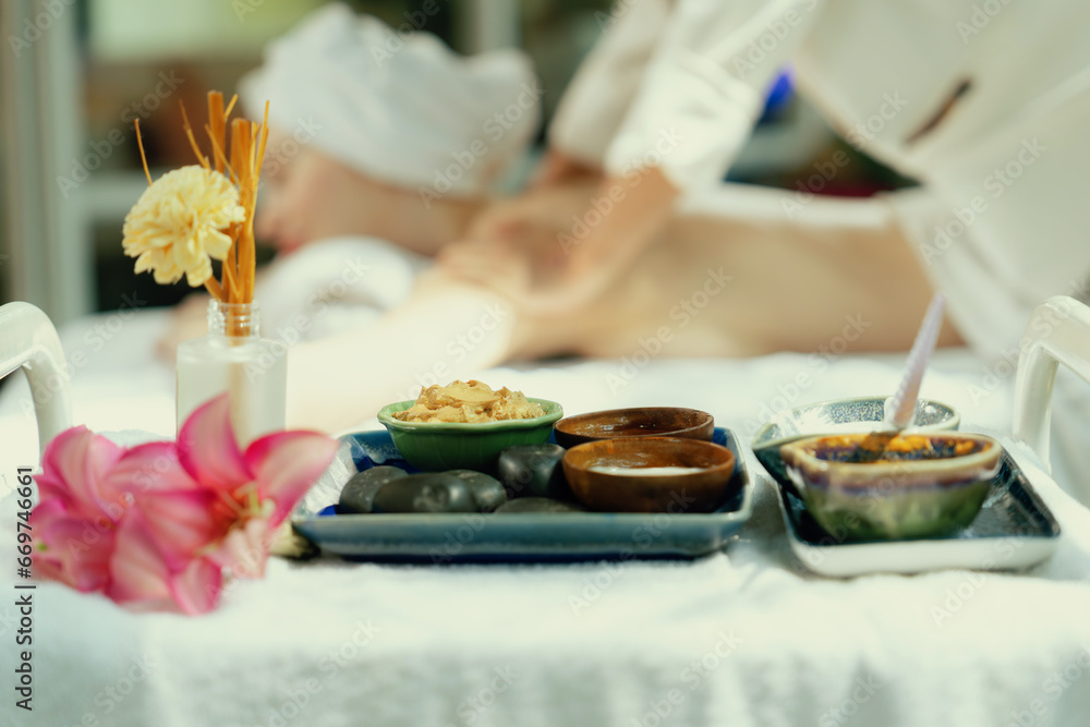 Natural herbal ingredient of facial mask and floral essence was placed on white towel in front of beautiful woman having body massage by professional masseuse. Blurring background. Tranquility.