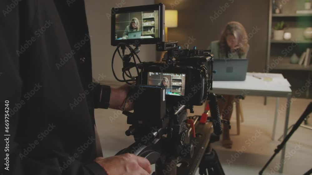 Medium tilt-down shot of male videographer setting up camera on tripod in studio, adjusting angle and frame, and female presenter sitting at table and using laptop, while preparing to film online show Medium tilt-down shot of male videographer setting up camera on tripod in studio, adjusting angle and frame, and female presenter sitting at table and using laptop, while preparing to film online show