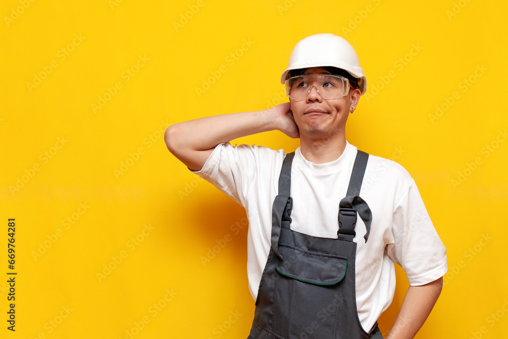 puzzled asian guy builder in uniform and safety glasses thinks and remembers on yellow isolated background
