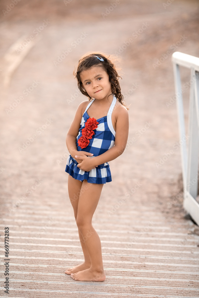 Little toddler girl in swimsuit standing at the beach and looking at camera, childhood memories and summer vacation concept Little toddler girl in swimsuit standing at the beach and looking at camera, childhood memories and summer vacation concept