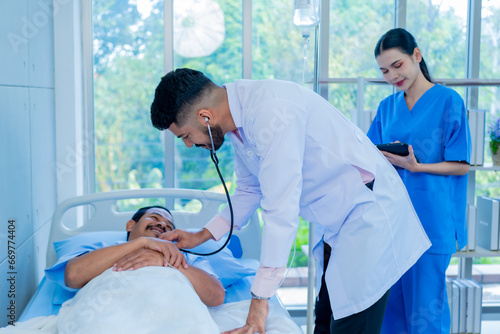 Asian professional male doctor in white lab coat using stethoscope listening monitoring heartbeat pulse on patient chest while female nurse in uniform writing symptom data on clipboard in ward room. photo