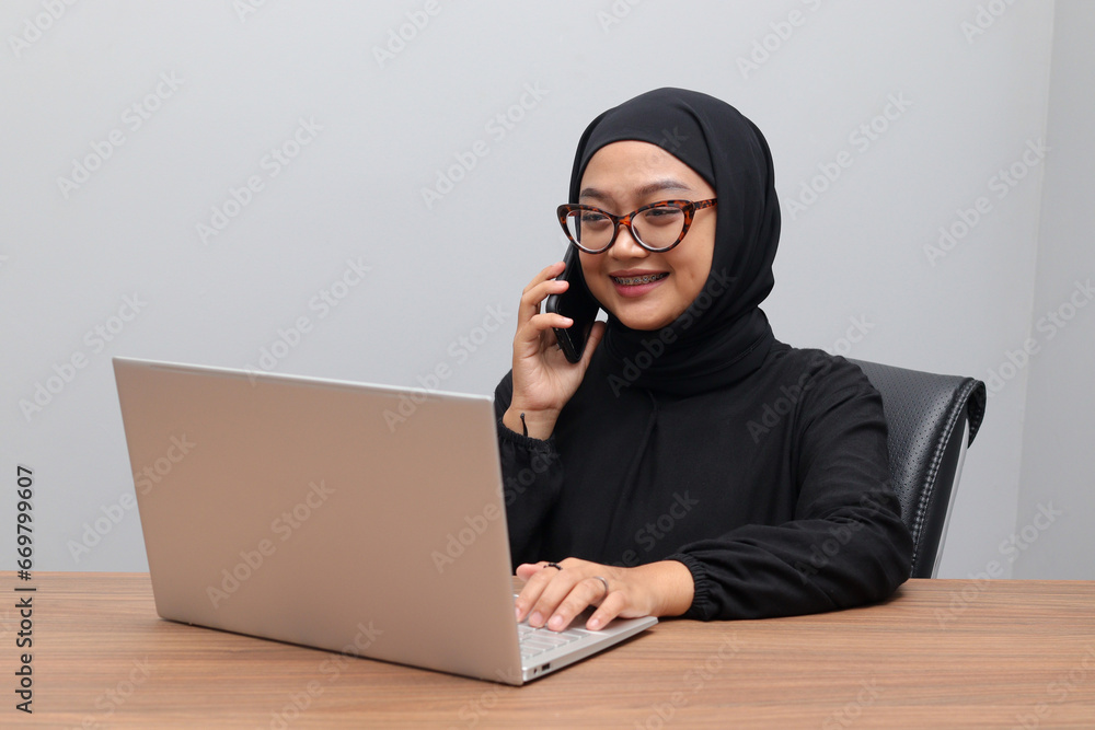 Portrait of attractive Asian hijab woman working on her laptop. Muslim girl making a phone call in office. Employee and freelance worker concept. Portrait of attractive Asian hijab woman working on her laptop. Muslim girl making a phone call in office. Employee and freelance worker concept.
