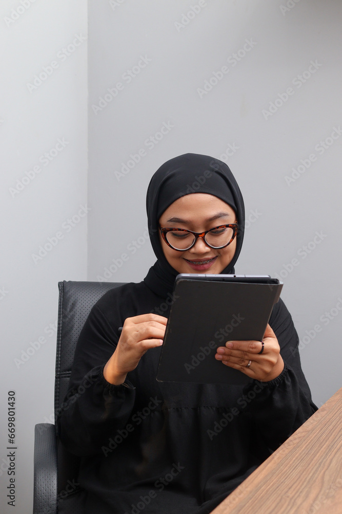 Portrait of attractive Asian hijab woman working on her tablet. Muslim girl doing task in office. Employee and freelance worker concept. Portrait of attractive Asian hijab woman working on her tablet. Muslim girl doing task in office. Employee and freelance worker concept.
