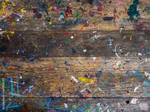 Wood plank top table covered in oil or acrylic color paint splatter. Abstract texture of old rustic wooden desk background of artist workshop studio with messy colorful color painted splash, top view.