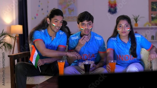 Indian cricket fans sitting on a couch and watching a live cricket match on TV - cricket team jersey. Friends sitting together in the living room with popcorn and cold drinks to watch Indian cricke... photo
