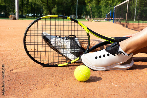 Close-up of tennis player's legs in sports shoes sneakers and a racket with yellow tennis ball. Female athlete sitting on the clay court next to the net. Sport victory tournament competition concept.
