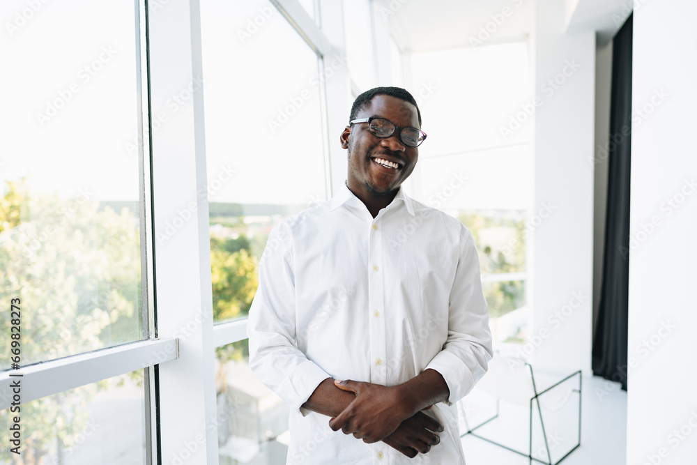 Fototapeta premium Young african man in white shirt and glasses posing near large window