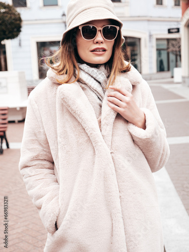 Young beautiful smiling lady wearing trendy white faux fur coat. Stylish woman posing in the street in winter. Cheerful and happy model in panama. Cold autumn weather. Shopping mall