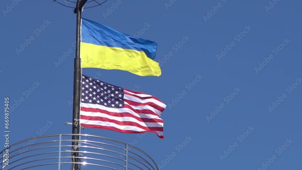 The flags of Ukraine and the USA flutter on a flagpole against a blue sky. State symbol. Ukrainian blue and yellow flag. Wind day outdoor. The flags of Ukraine and the USA flutter on a flagpole against a blue sky. State symbol. Ukrainian blue and yellow flag. Wind day outdoor.