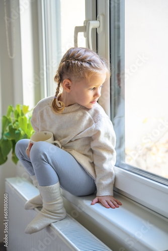 Little girl in warm sweater and socks sitting and looking out window, holding cup of hot drink and warming up from the heating radiator. Heating in an apartment, at home.