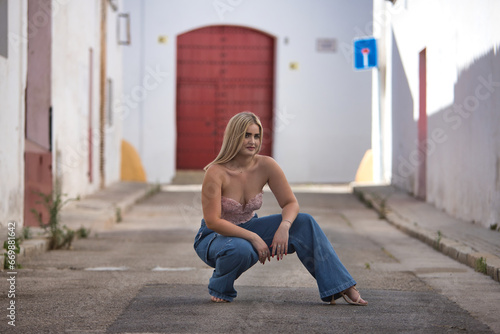 Young, blonde, green-eyed woman with pink top and jeans, posing crouched down on a lonely street in a pretty Andalusian village. Concept of beauty, places, villages, sunny.