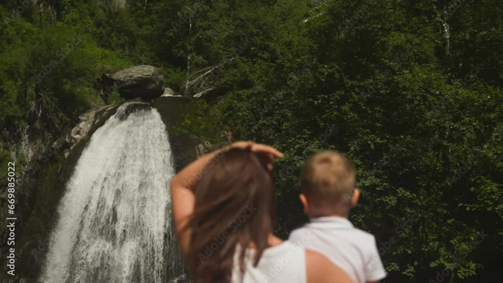 Mother adjusts long hair holding toddler son rest by waterfall in rocky park slow motion. Young woman shows with boy amazed by water cascade in wood Mother adjusts long hair holding toddler son rest by waterfall in rocky park slow motion. Young woman shows with boy amazed by water cascade in wood