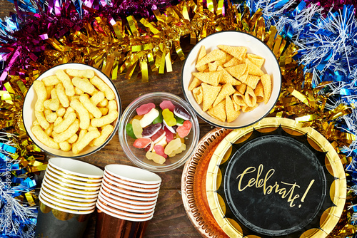 birthday or christmas, new year's party concept, paper plate with card celebrate text and disposable cup, straws and colorful with different chips and candies, pink hat tinsel on wooden table