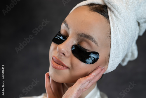 Beauty woman in towel applies black collagen patches under eyes isolated on black
