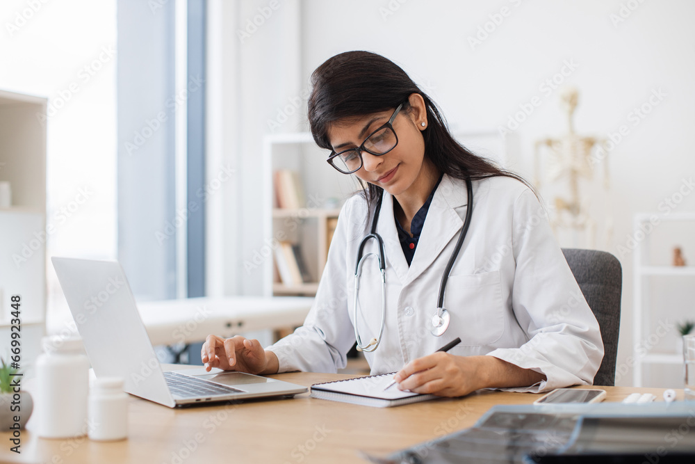 Side view of indian female doctor using portable computer while sitting at office desk in health center interior. Efficient mature general practitioner checking patient records via gadget. Side view of indian female doctor using portable computer while sitting at office desk in health center interior. Efficient mature general practitioner checking patient records via gadget.