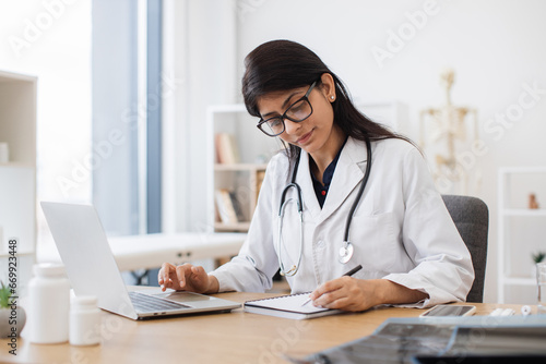Side view of indian female doctor using portable computer while sitting at office desk in health center interior. Efficient mature general practitioner checking patient records via gadget.