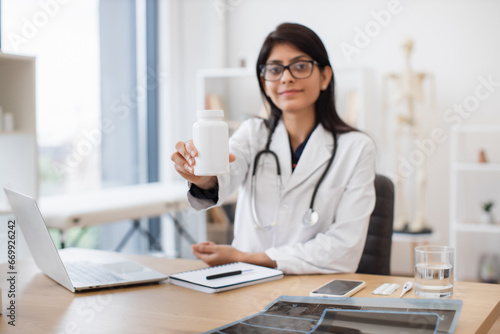 Thoughtful hindu female in medical white coat showing oral medication while sitting at office desk with laptop and notes. Family doctor giving recommendations about treatment therapy. © sofiko14