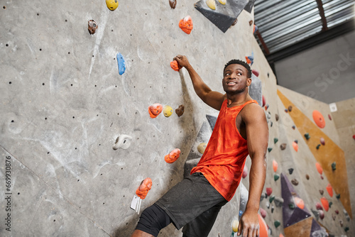 cheerful african american man in orange shirt ascending up rock wall and smiley looking away