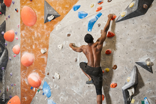 muscular shirtless african american man gripping strongly on boulders while climbing up rock wall