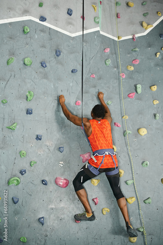 back view of african american man in orange shirt climbing actively up rock wall, bouldering concept